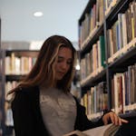 A young woman reading a book in a calm library setting, surrounded by shelves.