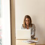 Businesswoman working remotely on a laptop at home, enjoying natural light in a modern, minimalist setting.