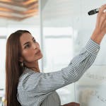 Woman in striped shirt presenting on a whiteboard in a modern office setting.